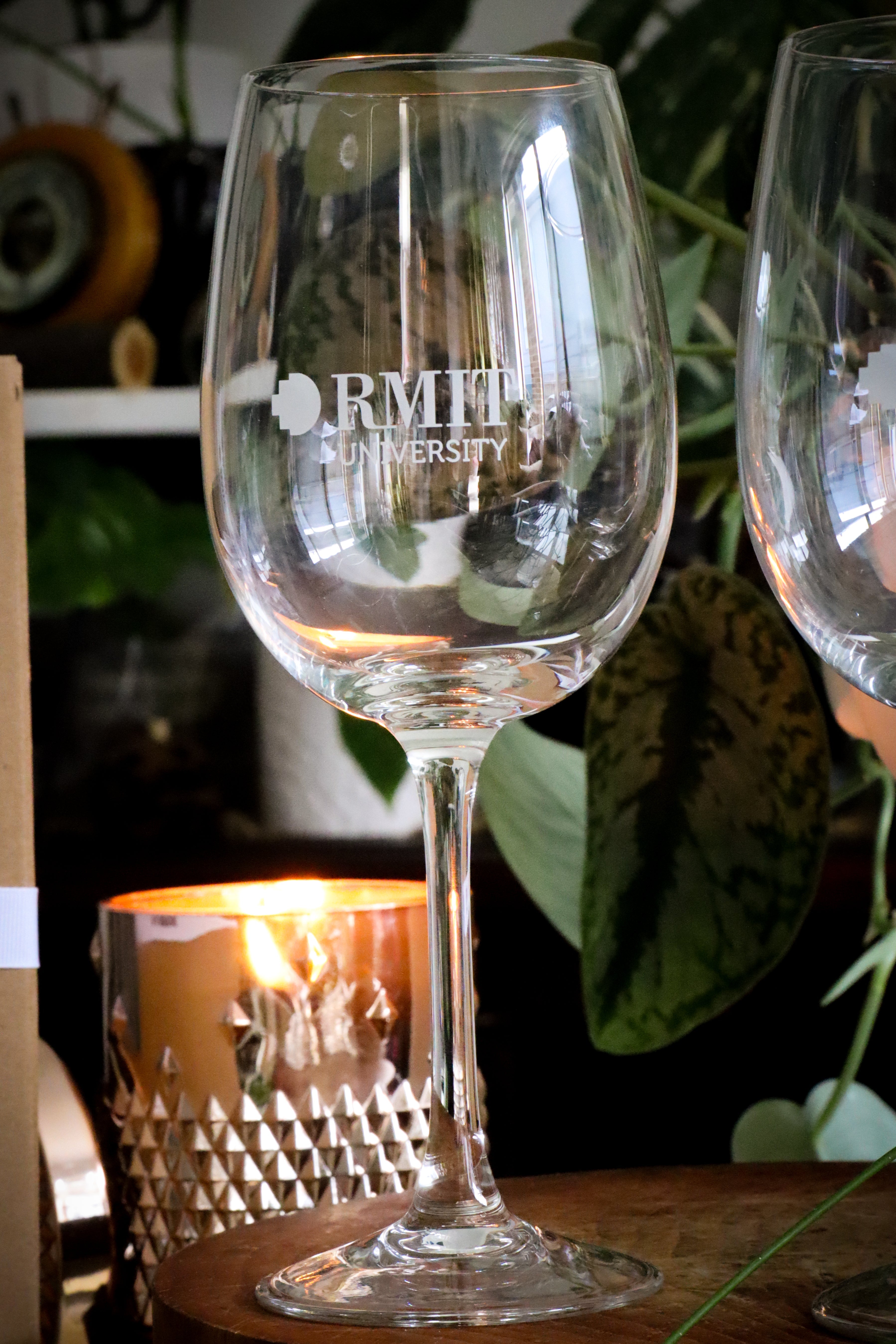 A close-up of a single wine glass etched with the RMIT University logo, showcasing the fine craftsmanship of the glass's curvature. In the background, a warm, lit candle and verdant foliage add to the serene and inviting setting. Available at the RMIT Store.